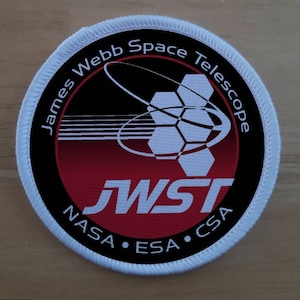 May include: A white circular patch with a black border and red and white design. The text "James Webb Space Telescope" is written in a circle around the design. The text "JWST" is written in white letters on a red background. The text "NASA • ESA • CSA" is written in a circle around the red background.