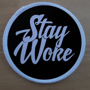 May include: A white embroidered patch with a black background and the text "Stay Woke" in a stylised font.