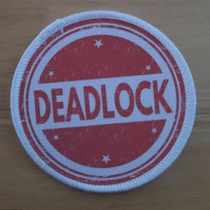 May include: White round patch with a red circle in the center. The red circle has a white border and the word "DEADLOCK" in white letters. The patch has a white stitched border.