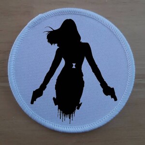 Könnte beinhalten: Runder Stoff-Patch in Weiß mit einer schwarzen Silhouette einer Frau, die zwei Pistolen hält. Die Frau trägt ein schwarzes Outfit mit einem Gürtel.