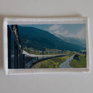 Puede incluir: Un tren azul viaja a través de un paisaje montañoso. El tren está en una vía que se curva alrededor de una ladera de la montaña. Las montañas están cubiertas de árboles verdes y hay una carretera junto a las vías.