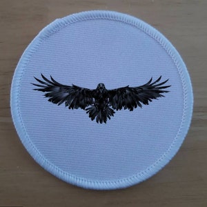 May include: A white round fabric patch with a black silhouette of a raven with its wings spread wide.
