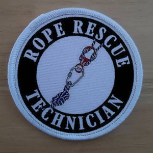 May include: White round patch with black border and text "ROPE RESCUE TECHNICIAN". A white background with a graphic of a rope and climbing gear is inside the circle.