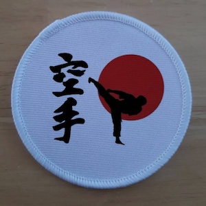 Puede incluir: Parche redondo blanco con un círculo rojo y una silueta negra de una persona pateando. La palabra japonesa "空手" está escrita en negro en el lado izquierdo del parche.