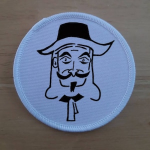 Op de afbeelding: Ronde witte stoffen patch met een zwart-witte illustratie van een man met een snor en een hoed. De man draagt een lange jas en heeft een lange baard.