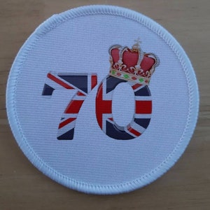 Könnte beinhalten: Ein weißer runder Patch mit einem rot-weiß-blauen Union-Jack-Design in Form der Zahl 70. Eine goldene Krone mit roten Juwelen sitzt über der Zahl.