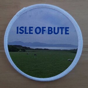 May include: A white round patch with a blue stitched border. The patch features a scenic image of a grassy field with a body of water in the background. The text "ISLE OF BUTE" is printed in blue letters at the top of the patch.