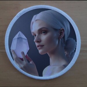 Pode incluir: Um remendo branco com uma borda redonda branca. O remendo apresenta uma mulher com cabelo branco segurando um grande cristal transparente na mão. O fundo é azul escuro.