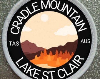May include: A black and white circular patch with the text "CRADLE MOUNTAIN LAKE ST CLAIR" around the edge. The text "TAS" appears on the left side and "AUS" appears on the right side. Inside the circle is a colorful illustration of a mountain range with a lake and trees in the foreground. The sky is a light orange color with a white sun and clouds.