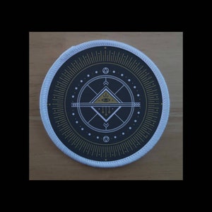 May include: Round patch with a white border and a black center featuring a geometric design. The design includes a triangle with an eye, lines, dots, and other geometric shapes in gold and white.