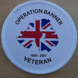 Puede incluir: Un parche redondo blanco con las palabras "OPERATION BANNER" en la parte superior. El parche presenta un mapa de Irlanda del Norte con el diseño de la bandera de la Union Jack. El texto "1969-2007 VETERAN" está impreso debajo.
