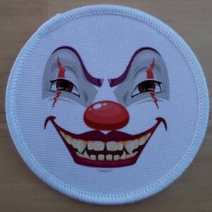 May include: A white round patch with a printed image of a clown's face. The clown has a red nose, a wide smile with sharp teeth, and red and purple makeup around the eyes.