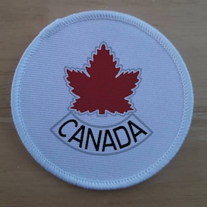 Peut inclure: Écusson rond en tissu blanc avec une feuille d'érable rouge et le mot "CANADA" dans une arche grise.
