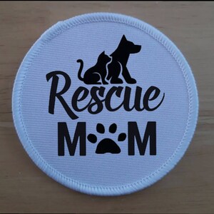 May include: A round, white patch with a stitched border. The patch features a black silhouette of a cat and dog, the words "Rescue Mom" in a script font, and a paw print. The design is centered on the patch.