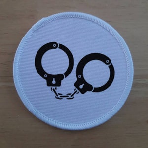 May include: A white fabric patch with a black silhouette of handcuffs. The handcuffs are linked together with a chain.