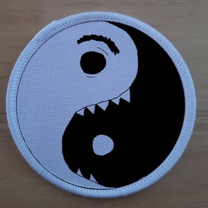 Può includere: Un simbolo yin yang in bianco e nero con un viso da cartone animato e denti sul lato bianco. Il simbolo è su una toppa di tessuto bianco con un bordo bianco cucito.