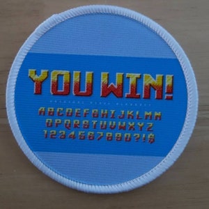 May include: A round, white patch with a blue background. The text "YOU WIN!" is in yellow and red pixelated letters. Below the text is a pixelated alphabet and numbers in yellow and red.