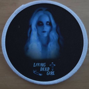 Pode incluir: Um remendo redondo branco com fundo preto. O remendo apresenta uma imagem azul de uma mulher com cabelo comprido e uma cruz. O texto "LIVING DEAD GIRL" é impresso abaixo da imagem.