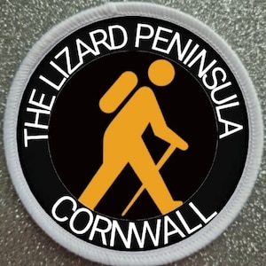 Pode incluir: Um remendo redondo branco com texto preto e branco que diz "The Lizard Peninsula Cornwall". Um círculo preto com uma figura de pau de caminhante amarela está no centro.