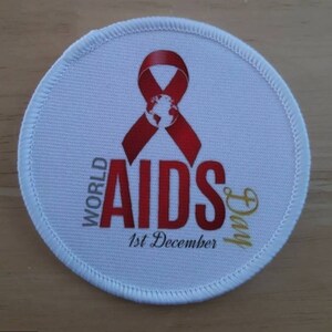 Puede incluir: Un parche redondo blanco con una cinta roja y las palabras "WORLD AIDS Day 1st December" en letras rojas y doradas. La cinta tiene un globo en el centro. El parche tiene un borde blanco cosido.