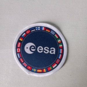 Könnte beinhalten: Ein runder, blauer Patch mit dem Text "e esa" und einem Kreis mit einem Punkt in der Mitte. Der Patch ist von einem weißen Rand mit kleinen Flaggen verschiedener Länder umgeben.