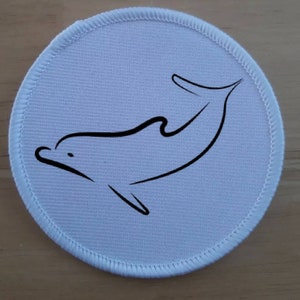 May include: White round fabric patch with a black outline of a dolphin jumping out of the water.