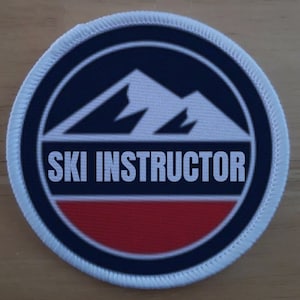 Op de afbeelding: Een ronde witte patch met een blauwe rand en een wit bergketenontwerp. De tekst "SKI INSTRUCTOR" is in zwart onder de bergen gedrukt. De onderkant van de patch is rood.