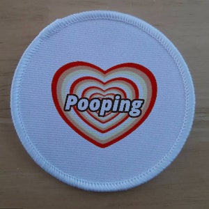 Op de afbeelding: Ronde witte stoffen patch met een rood hartontwerp. Het hart heeft concentrische cirkels van beige en wit. De tekst "Pooping" staat in zwarte letters in het hart.