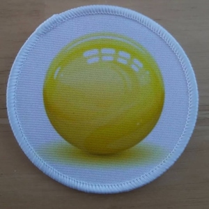 Puede incluir: Un parche redondo de tela blanca con una esfera amarilla en el centro. La esfera tiene una textura sutil y una ligera sombra debajo.