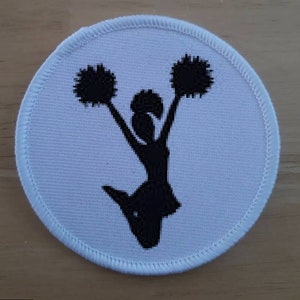 Può includere: Toppa rotonda bianca con una silhouette nera di una cheerleader che tiene pompon.