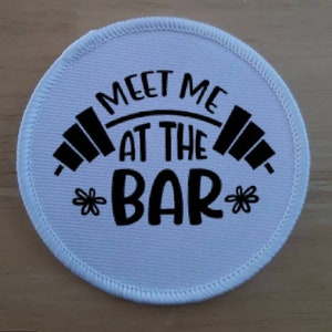 May include: White round fabric patch with black text that reads "Meet me at the bar" with a barbell graphic above the text and a flower graphic on either side of the text.