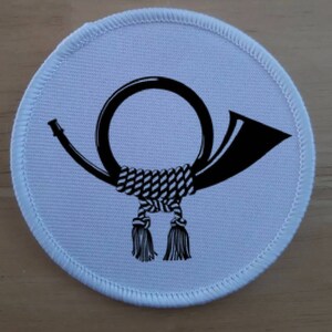 May include: A white round patch with a black and white image of a postal horn. The horn is tied with a rope and has tassels.