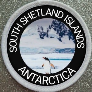 May include: A white round patch with a black border and the text "SOUTH SHETLAND ISLANDS ANTARCTICA" around the edge. The centre of the patch features a photo of a penguin standing on a snowy landscape with mountains in the background.