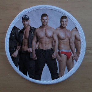 Peut inclure: Un patch circulaire blanc avec une bordure blanche et trois hommes torse nu. Les hommes portent un pantalon noir, une veste noire et des sous-vêtements rouges. Les hommes sourient et regardent la caméra.