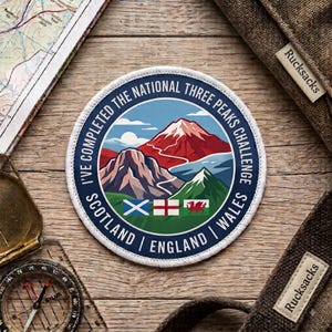 Może przedstawiać: Okrągła haftowana naszywka przedstawiająca górski krajobraz z napisem I'VE COMPLETED THE NATIONAL THREE PEAKS CHALLENGE SCOTLAND ENGLAND WALES. Wzór zawiera flagi Szkocji, Anglii i Walii umieszczone pod trzema szczytami górskimi.