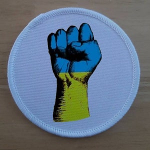 May include: A white circular patch with a raised edge, featuring a stylized image of a raised fist painted in the colors of the Ukrainian flag: blue and yellow.