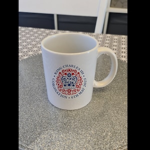 Könnte beinhalten: Weiße Keramiktasse mit einem blauen und roten kreisförmigen Design mit einer Krone und dem Text "King Charles III Coronation 6th May 2023".