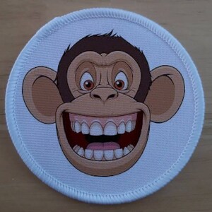 May include: A white round patch featuring a cartoon image of a monkey with a wide grin and large teeth. The monkey has brown fur and a white face.
