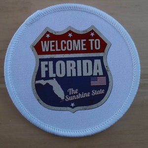 May include: White round patch with a blue and red vintage-style road sign that says "Welcome to Florida" with a map of Florida and the text "The Sunshine State".