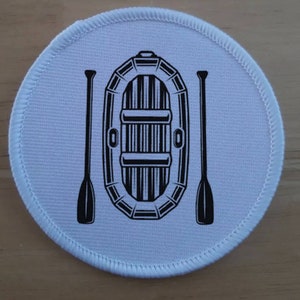 Puede incluir: Un parche redondo blanco con una ilustración en blanco y negro de un pequeño bote inflable con dos remos.