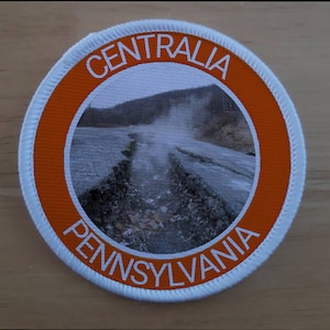 Könnte beinhalten: Ein runder weißer Patch mit orangefarbenem Rand. Der Patch zeigt ein Bild einer dampfenden Landschaft mit dem Text "CENTRALIA PENNSYLVANIA" um das Bild herum.