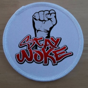 May include: A circular white patch featuring a black raised fist and the red text "STAY WOKE", outlined in black. The patch has a white stitched border.
