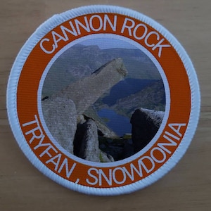May include: A white and orange round patch with the text "Cannon Rock Tryfan, Snowdonia" around the edge. The center of the patch features a photo of a rocky mountain landscape with a lake.