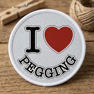 May include: A circular embroidered patch with a light gray background. The design features a large black letter I, a solid red heart symbol, and the word PEGGING in bold black capital letters arranged in a curved arc along the bottom edge.