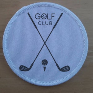 May include: A white, circular patch with a stitched border. The patch features a black graphic of crossed golf clubs, a golf ball, and the words "GOLF CLUB" in a sans-serif font. The design is centered on the patch.