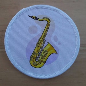 May include: A white round patch with a gold saxophone on it. The saxophone is a musical instrument with a curved neck and a bell at the end.