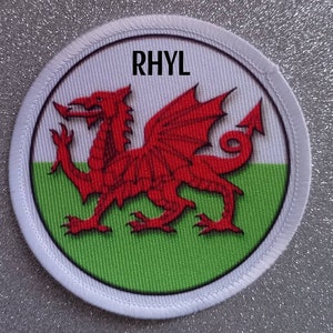Peut inclure: Un écusson circulaire blanc avec un dragon rouge sur un fond vert et blanc. Le texte "RHYL" est imprimé en noir en haut de l'écusson.
