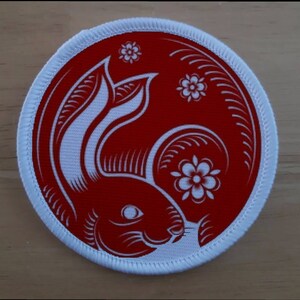 May include: A white and red circular patch with a white rabbit design and white flowers. The rabbit is in a curled position with its head facing the right side of the patch. The background is red.