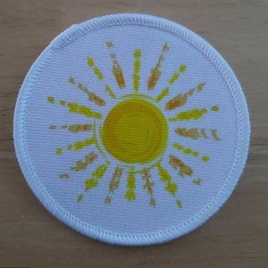 May include: A round, white patch with a yellow sun design. The sun has a yellow center and orange rays. The patch has a white stitched border.