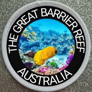 Op de afbeelding: Een ronde geborduurde patch met een levendige onderwaterscène van het Great Barrier Reef in Australië. Het midden toont gele koraalvlinders, koraalformaties en kleinere vissen. De zwarte rand bevat de witte tekst THE GREAT BARRIER REEF AUSTRALIA.
